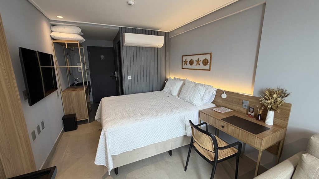 Visão geral do flat: cama, bancada de trabalho com cadeira de palha, televisão e armário em madeira